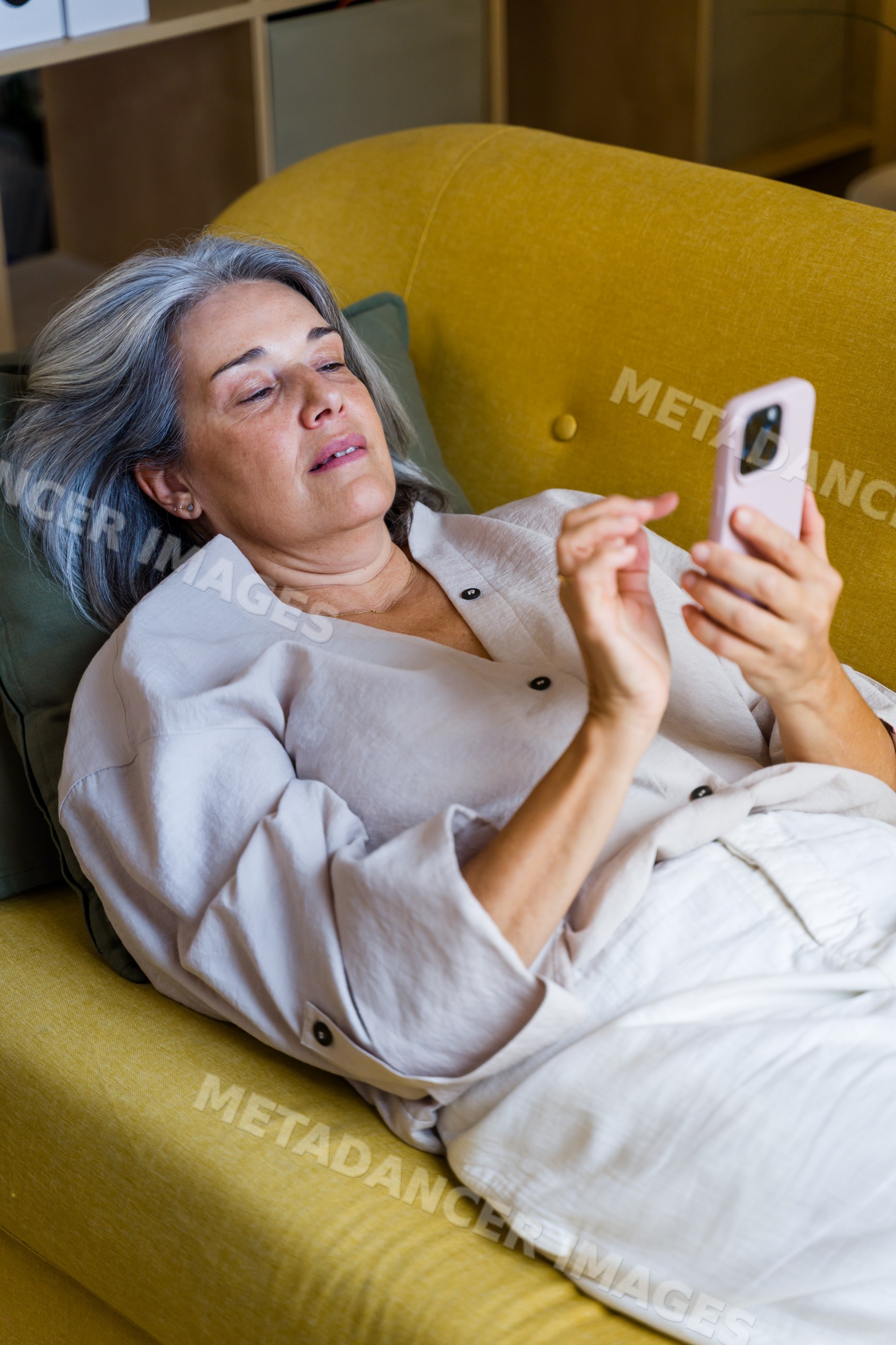 White woman lying on yellow couch using smartphone indoors