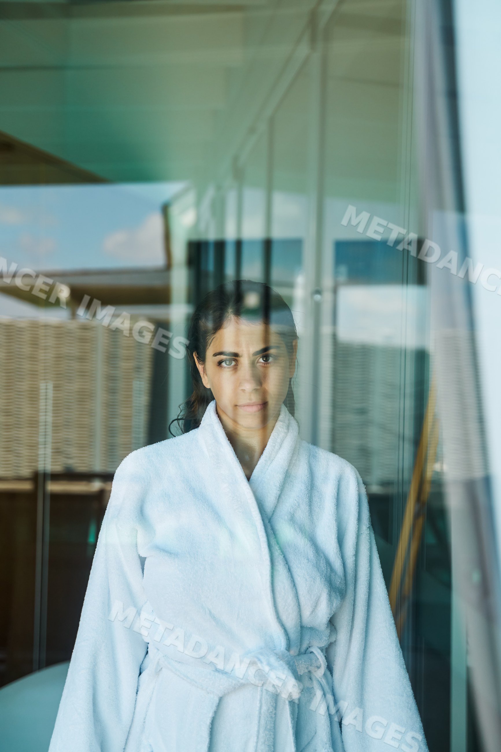 A woman in a white bathrobe stands indoors near a glass window