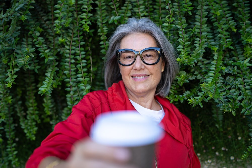 Mature white woman holding coffee cup outdoors with green foliage