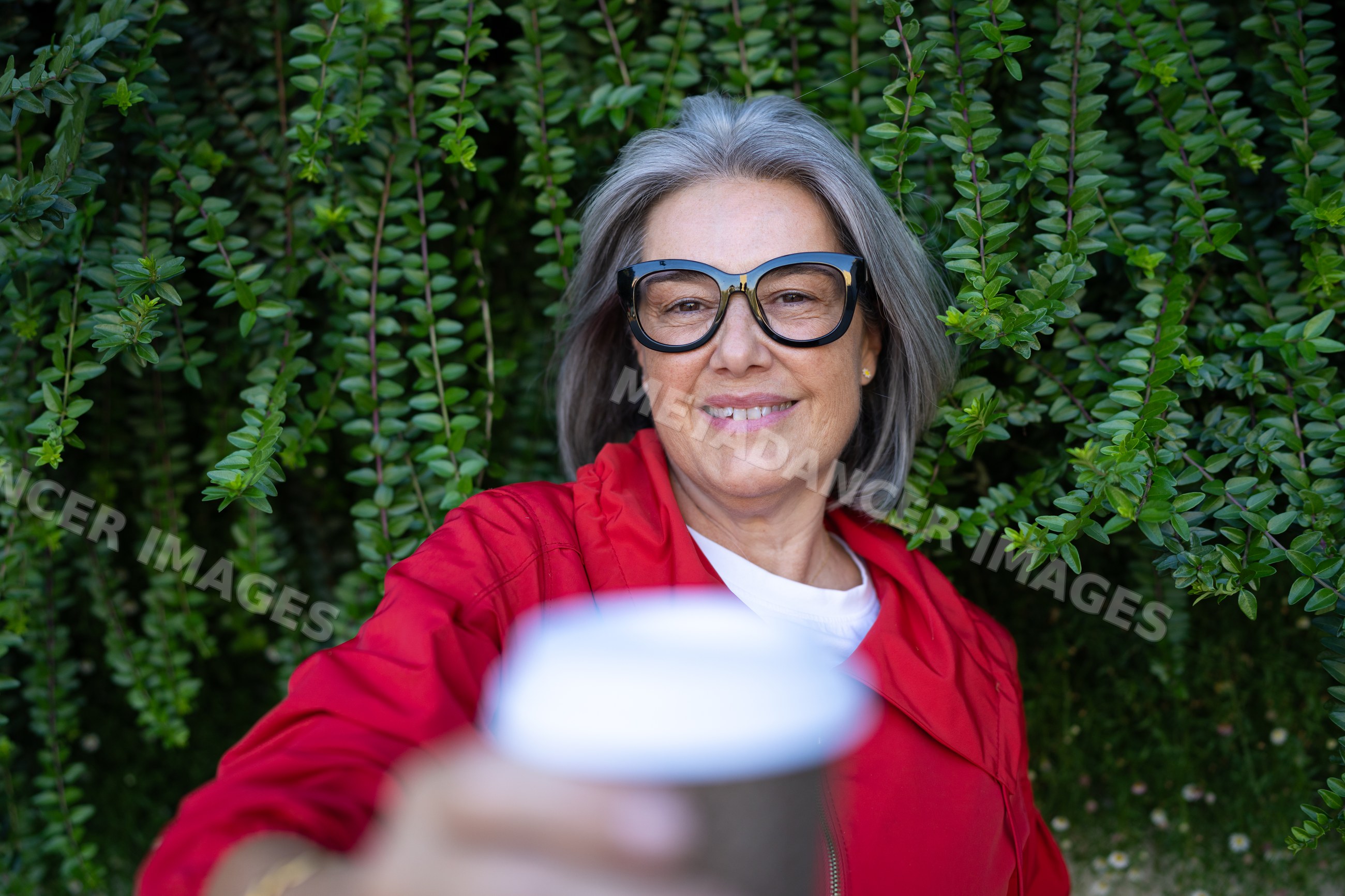Mature white woman holding coffee cup outdoors with green foliage
