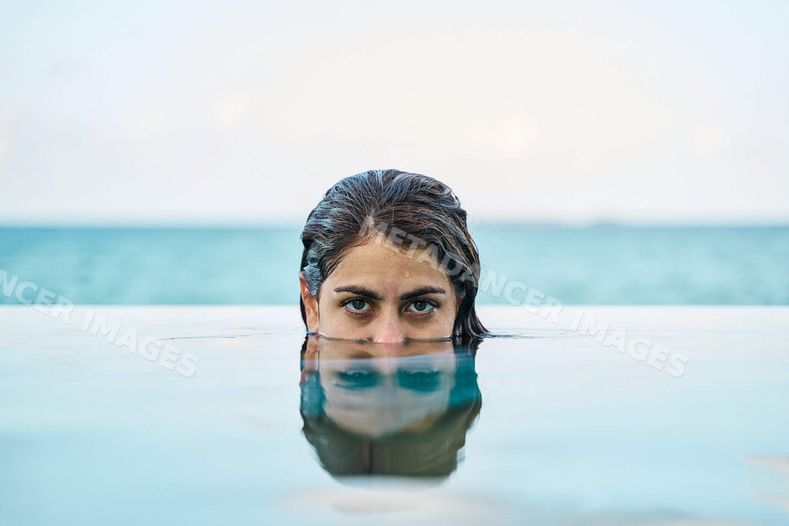 A white woman partially submerged in an outdoor pool with ocean view