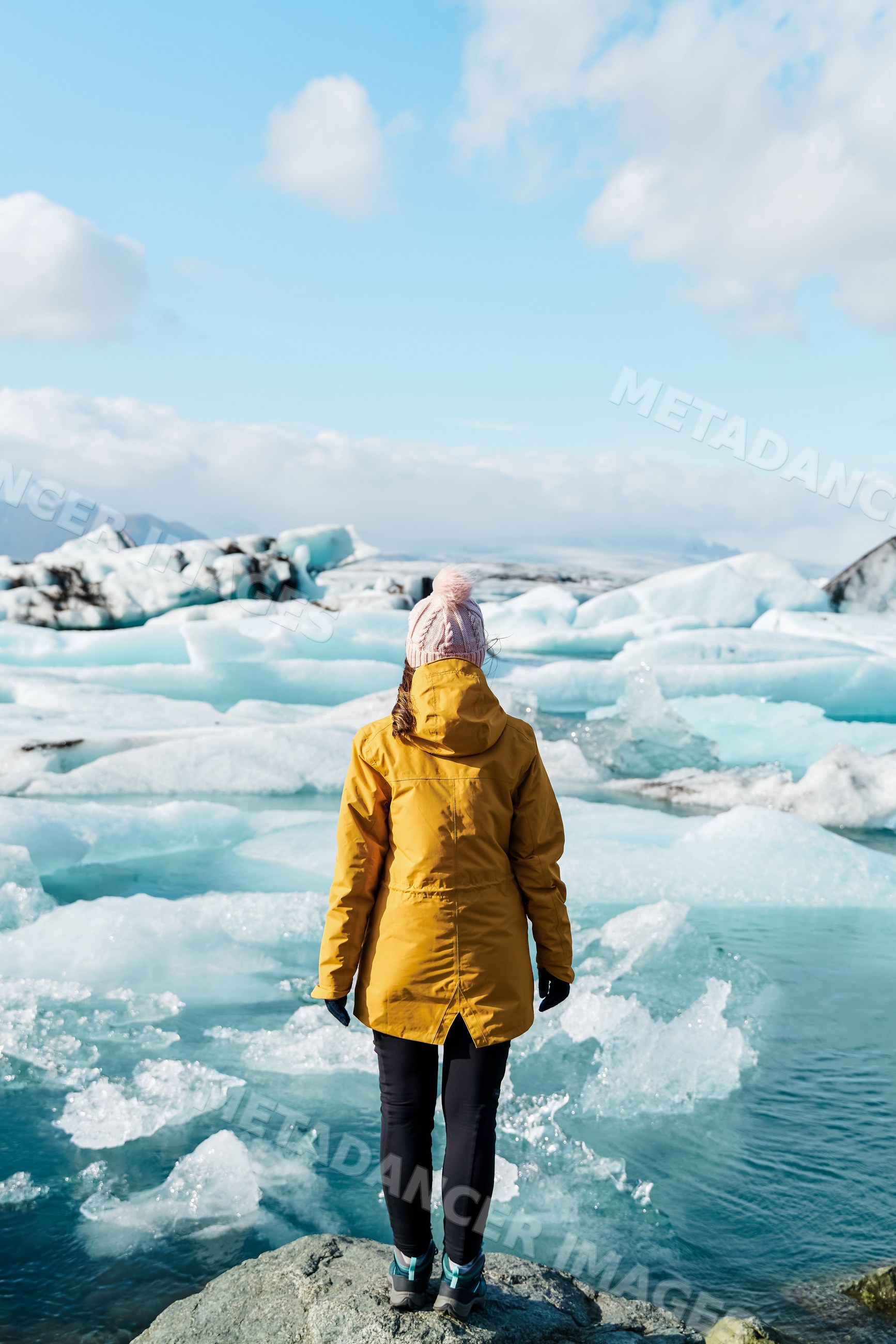 A white woman in winter clothing stands on a rock looking at icebergs
