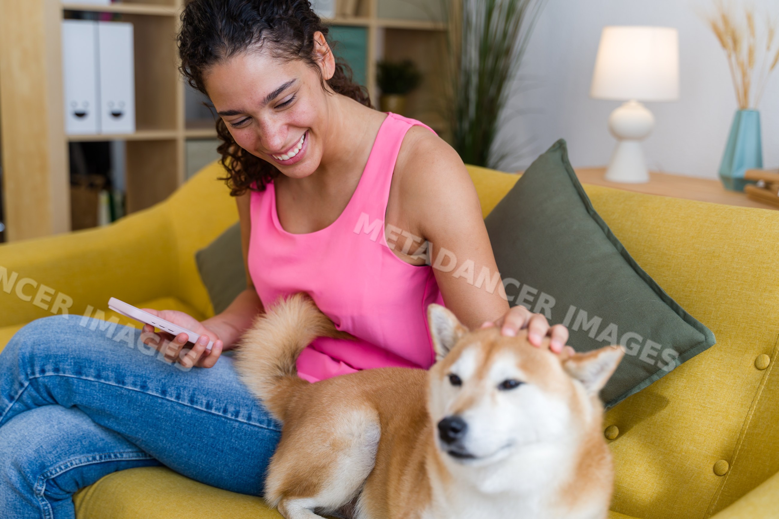 A smiling Hispanic woman pets a small dog while holding a phone