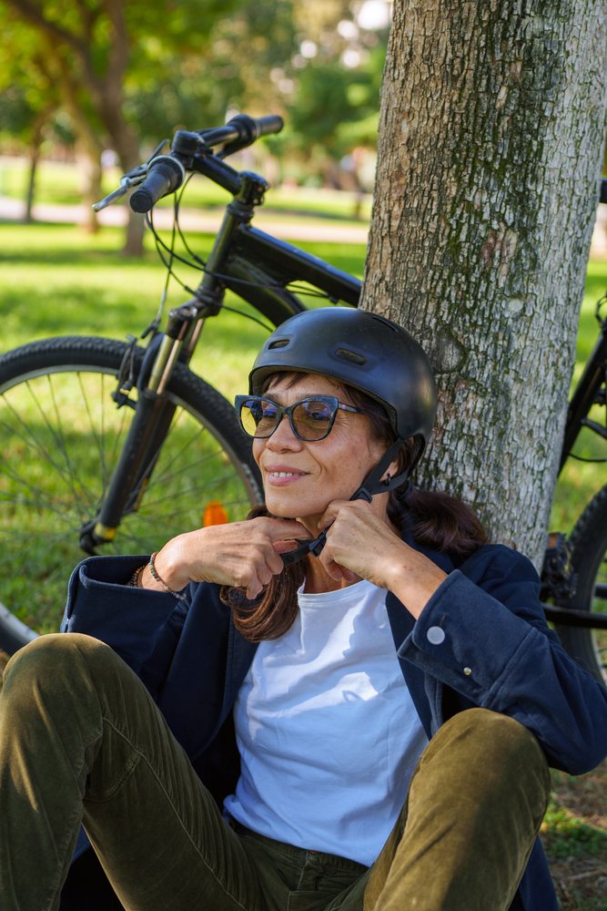 Hispanic woman adjusting helmet strap resting by bicycle under tree