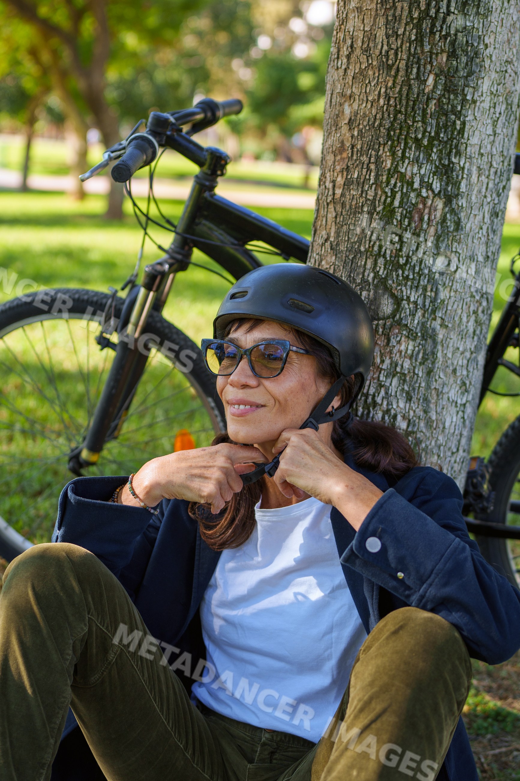 Hispanic woman adjusting helmet strap resting by bicycle under tree