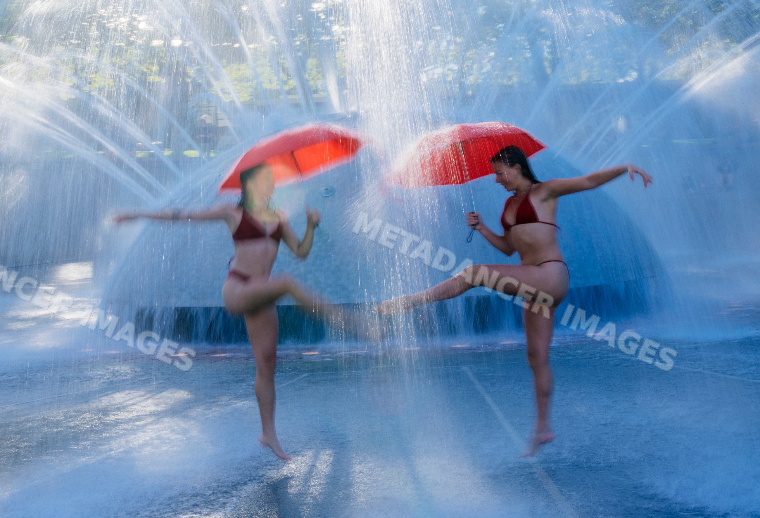 Two white women dancing with red umbrellas in water fountain in USA