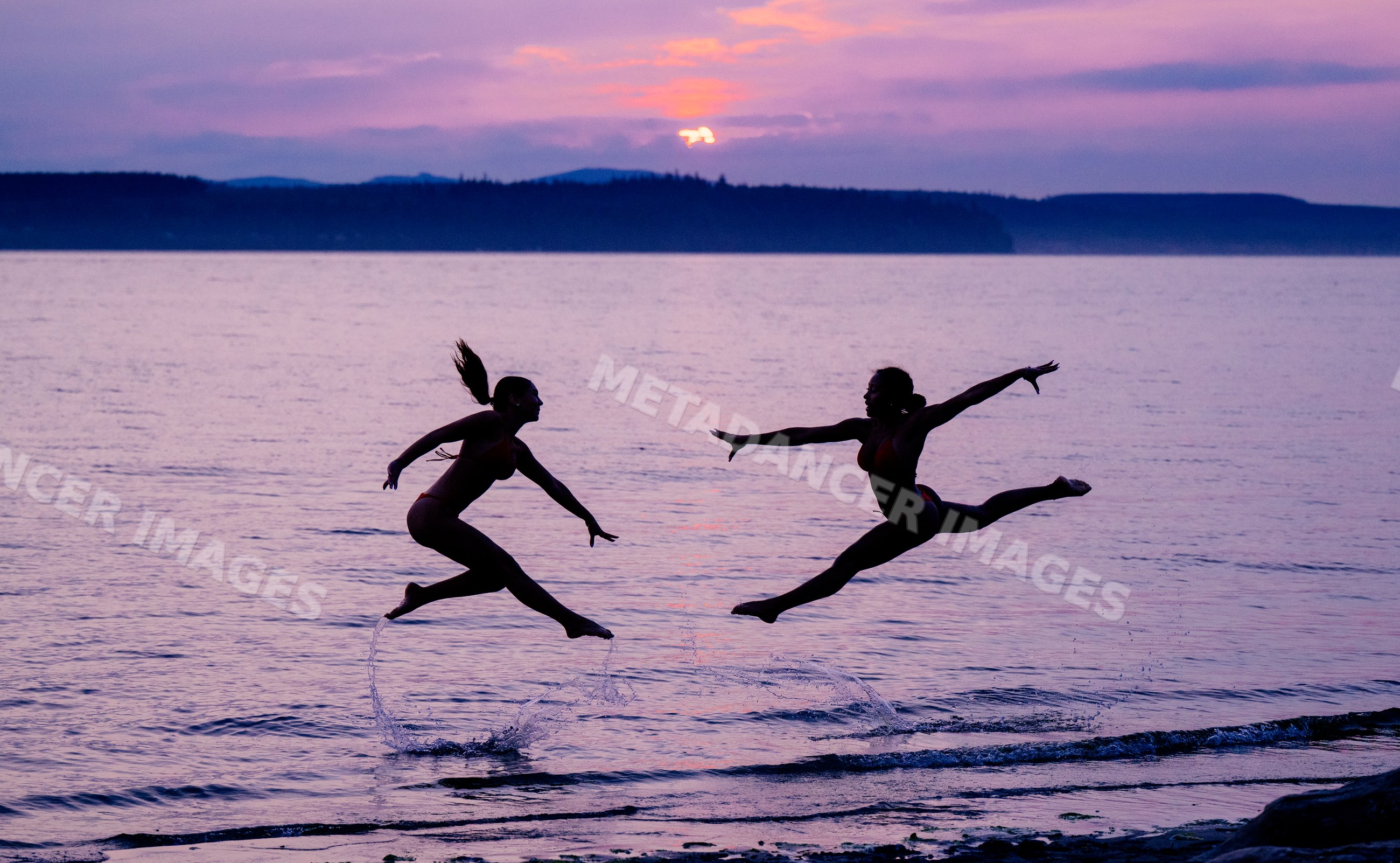 Two women perform acrobatic jumps in water at sunset on lake shore