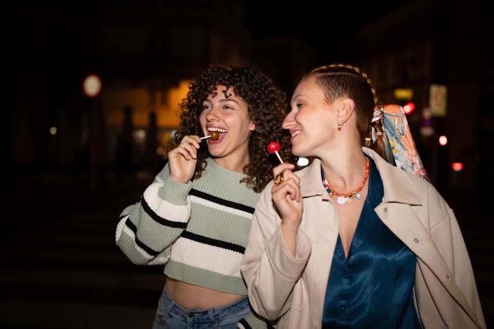 Two white women enjoying lollipops together at night in the city