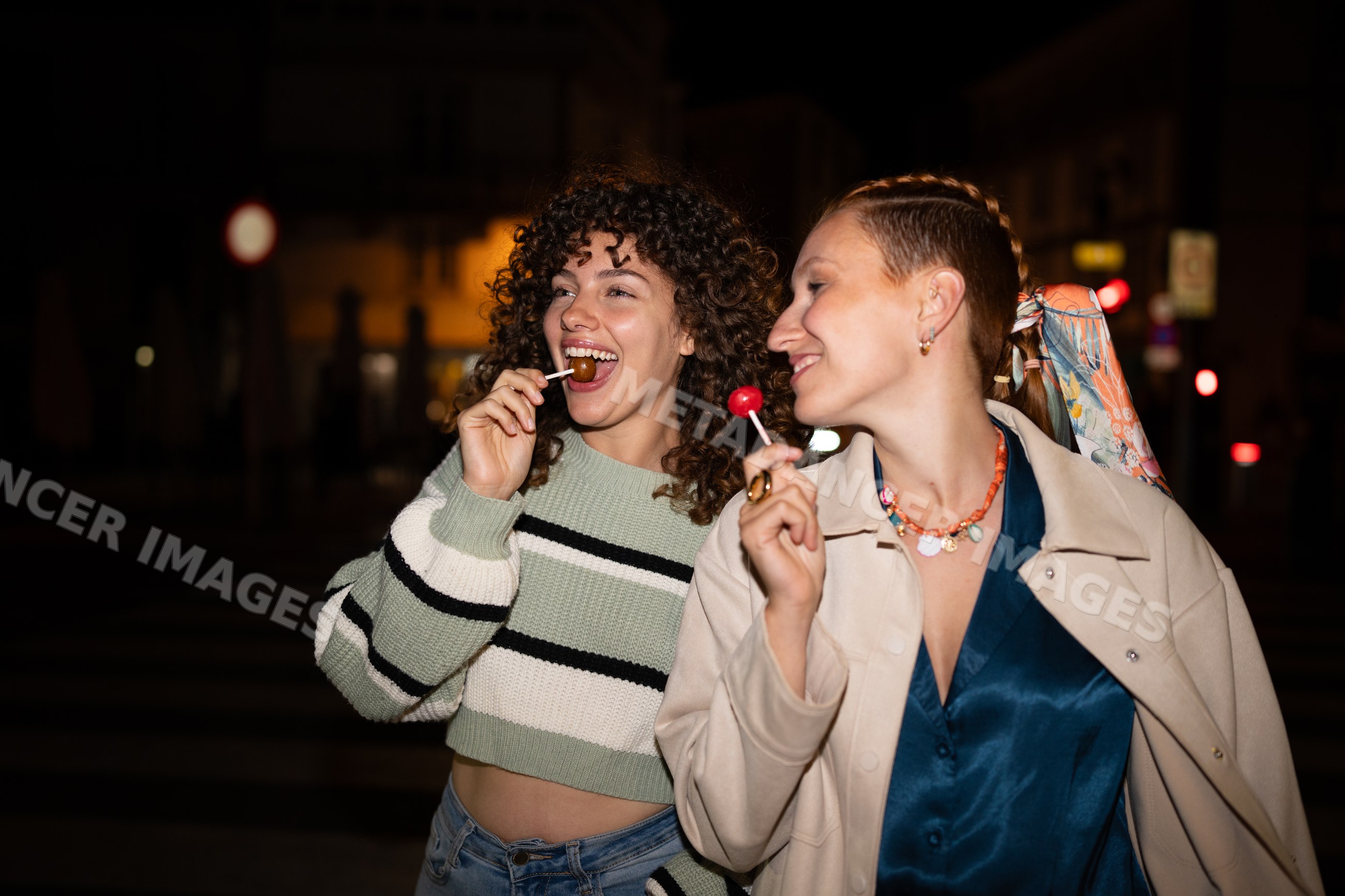 Two white women enjoying lollipops together at night in the city