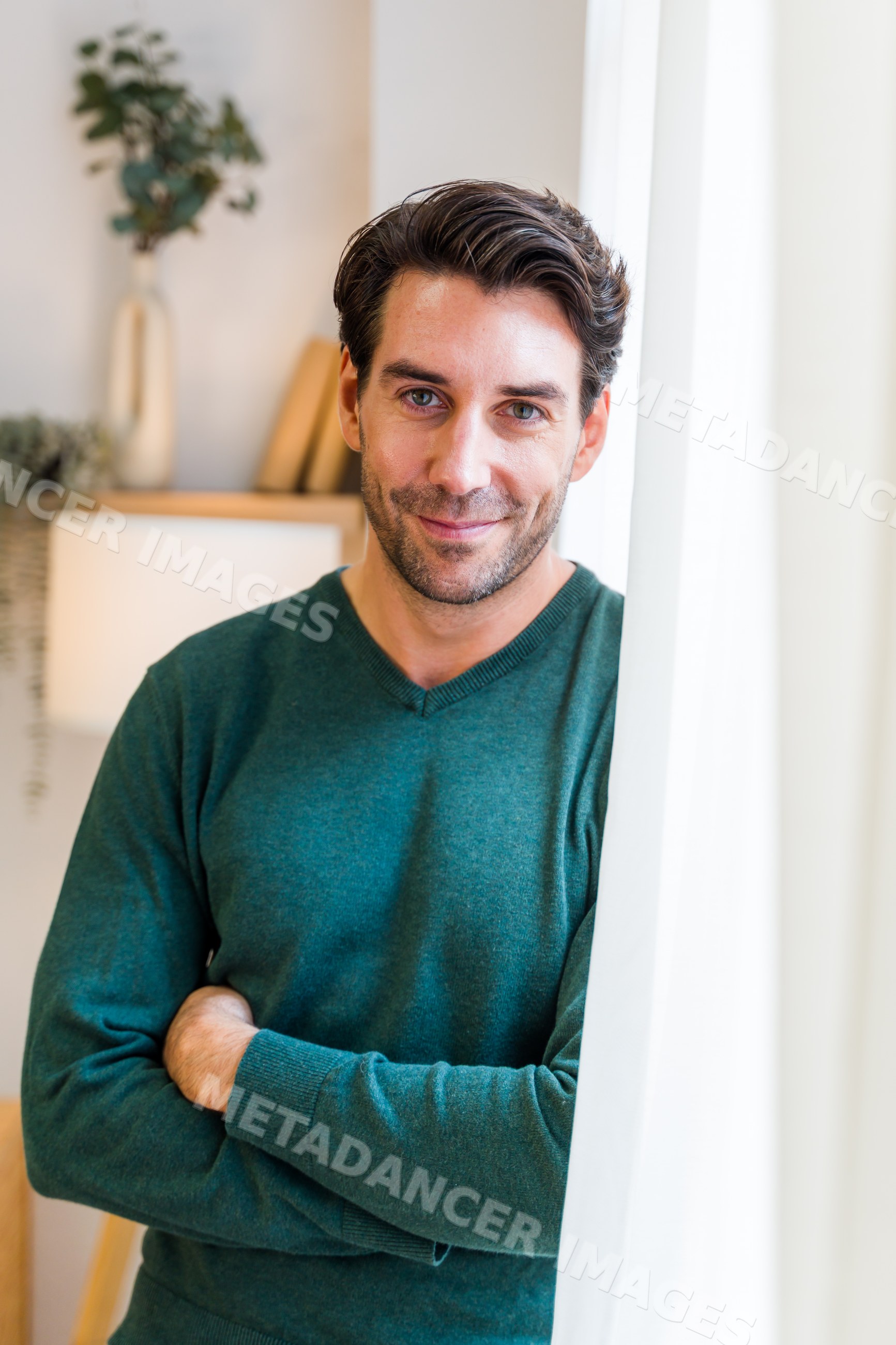 White man in green sweater standing indoors beside curtain