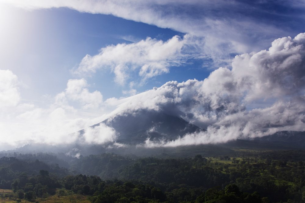 Volcanic mountain rising above forest with cloud cover and blue sky