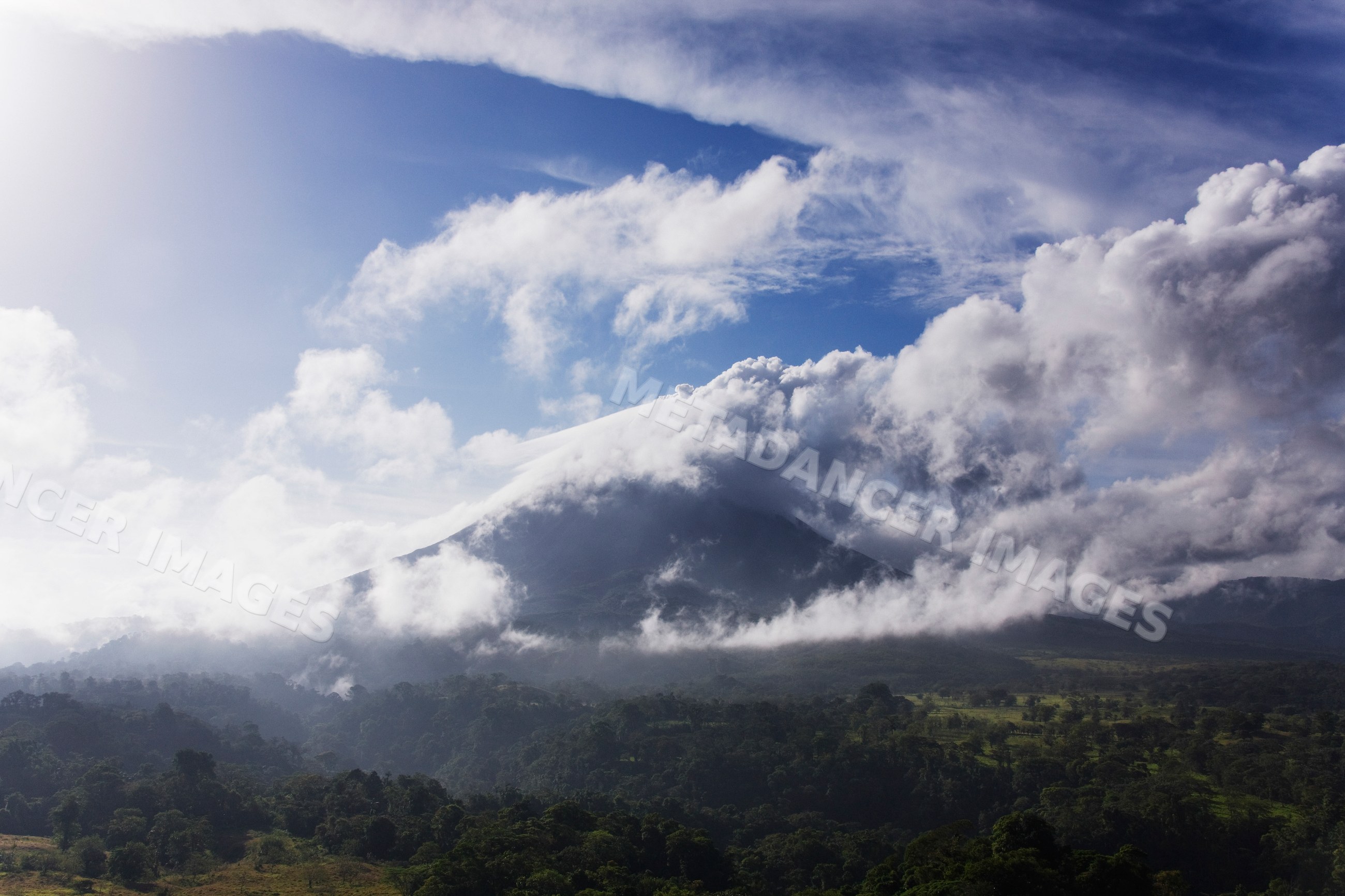Volcanic mountain rising above forest with cloud cover and blue sky