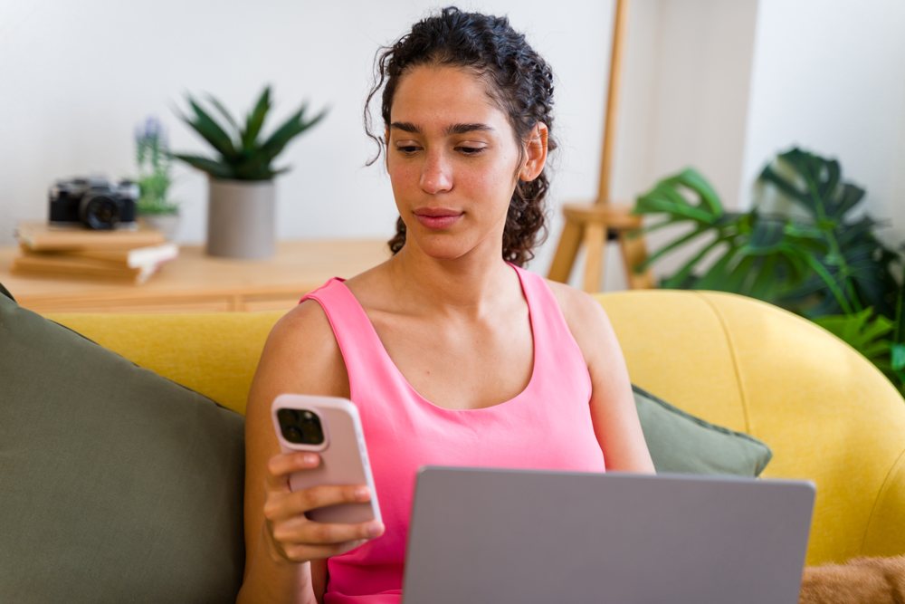 A Hispanic woman multitasks using phone and laptop at home in Spain