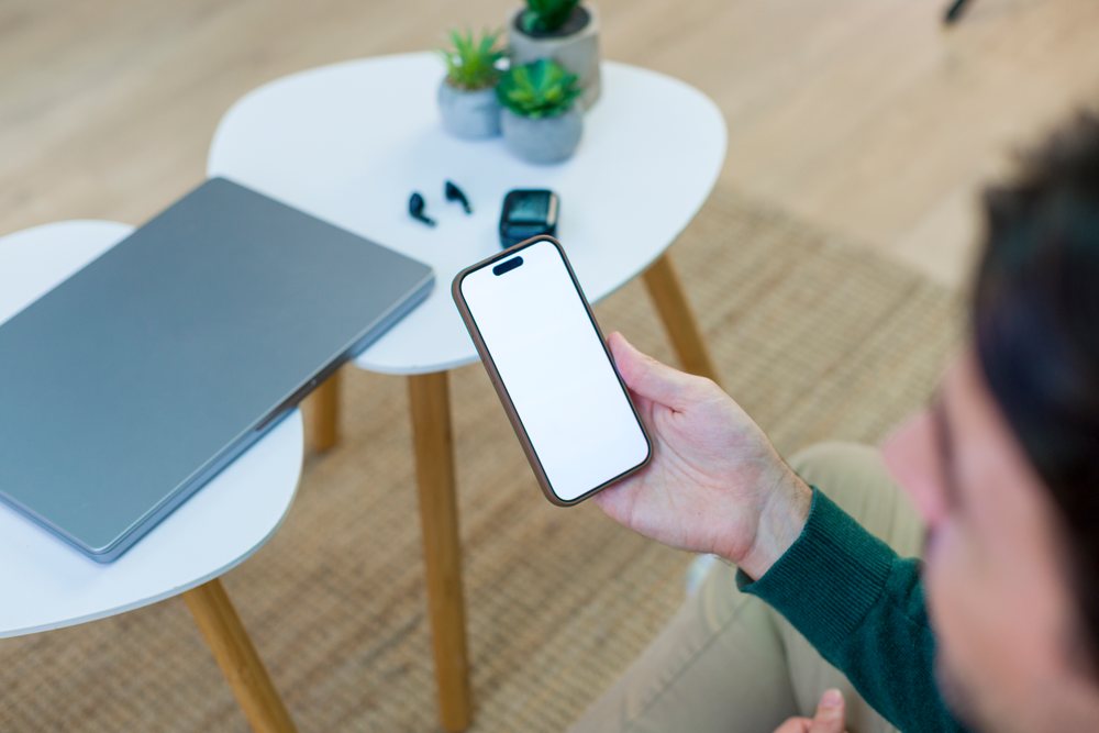 White person holding smartphone by table with laptop, earbuds, watch