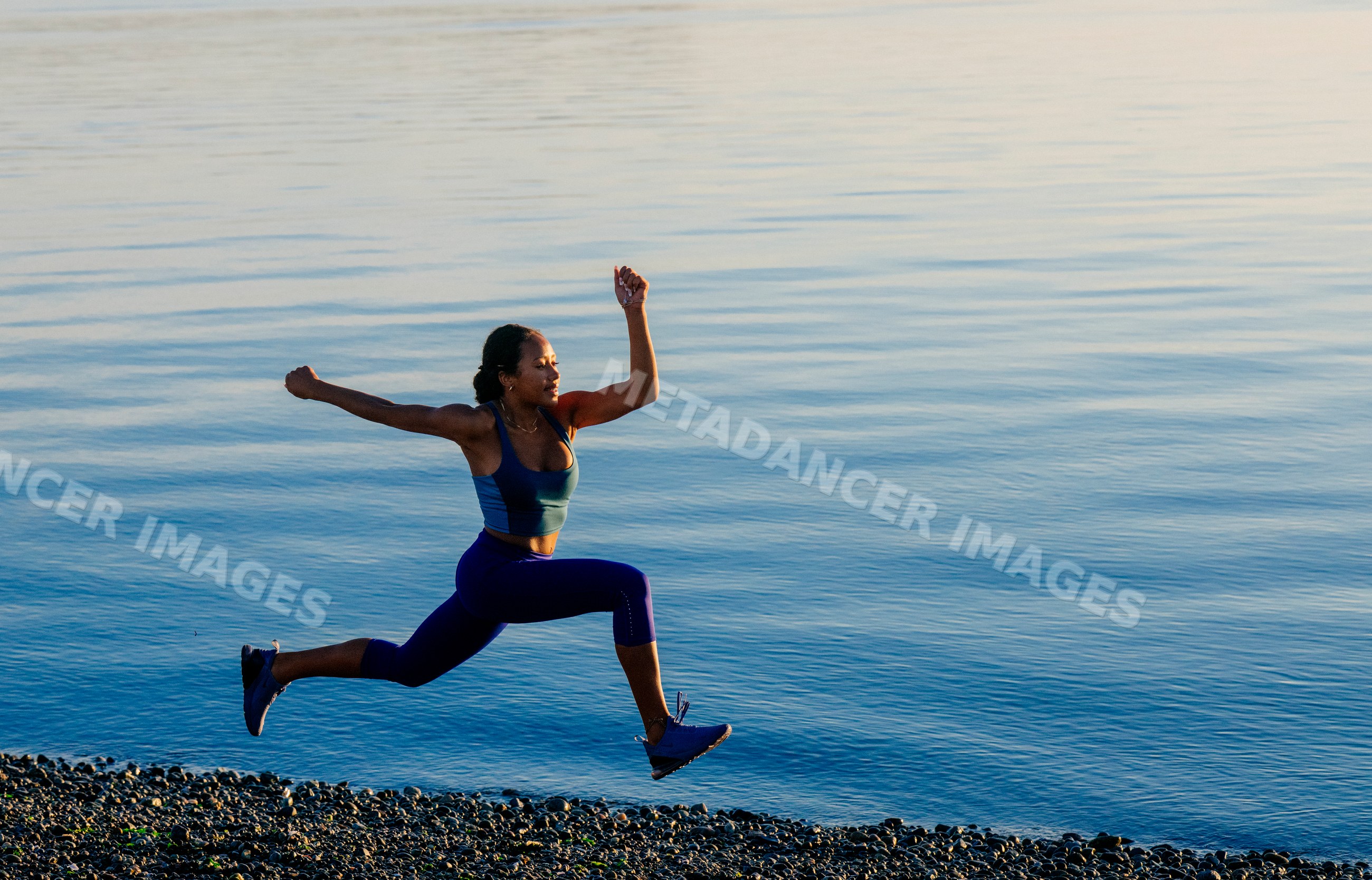 Hispanic woman jumping along pebbled shoreline by calm lake