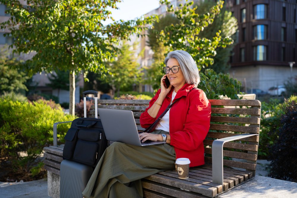 White woman sitting on bench using laptop and phone outdoors
