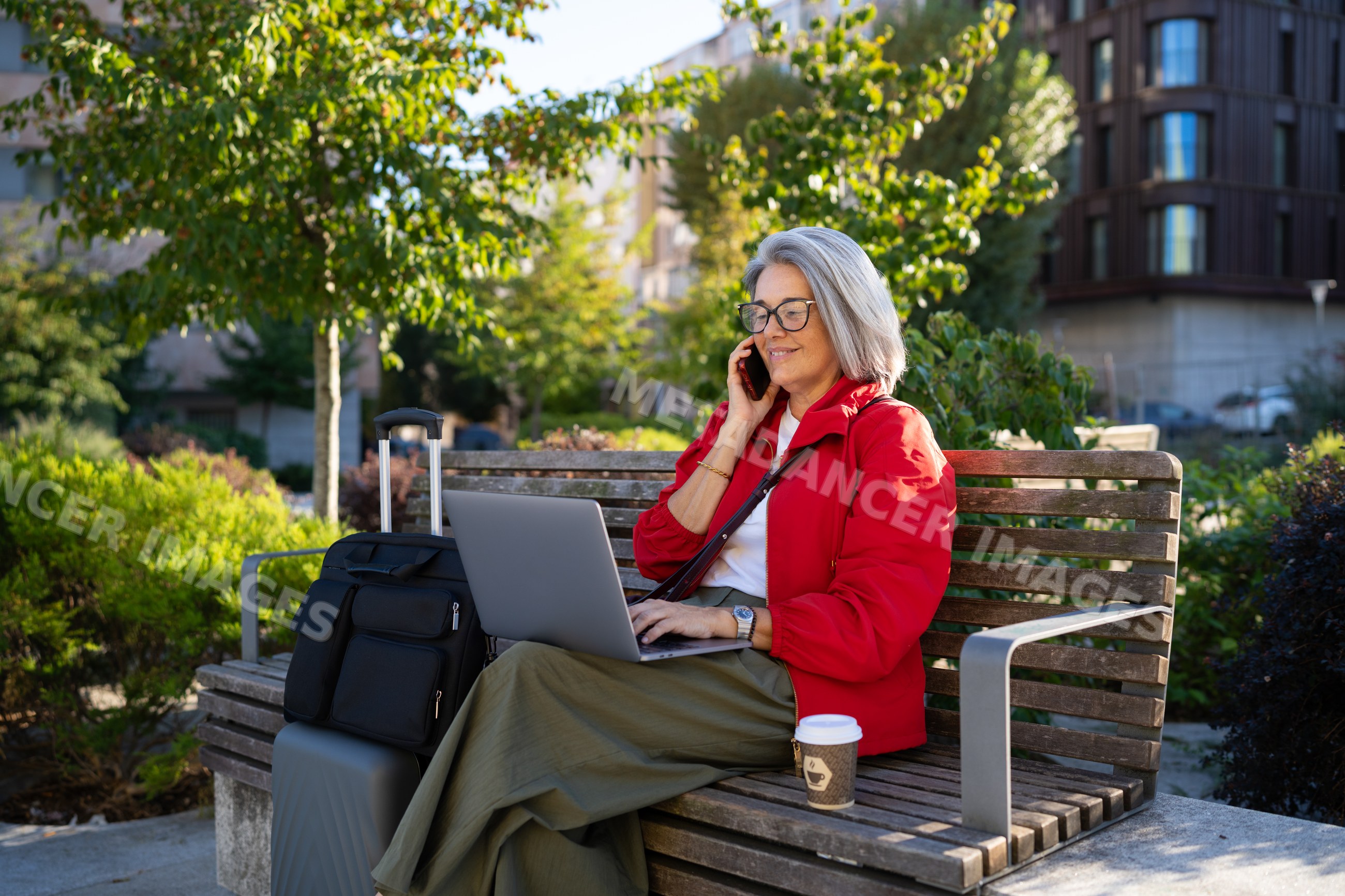 White woman sitting on bench using laptop and phone outdoors