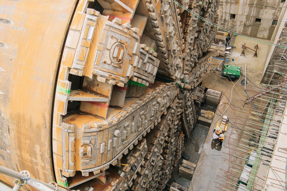 Two white construction workers inspect plans beside a large tunnel