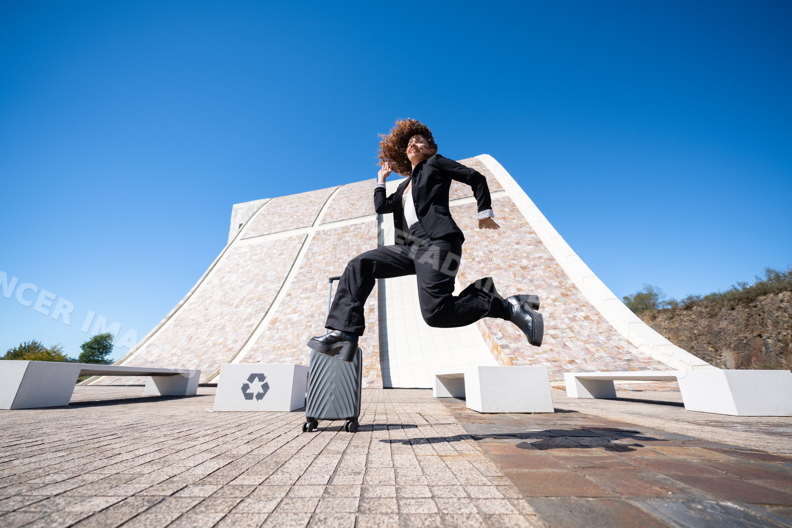White woman jumping with suitcase in urban plaza in Spain