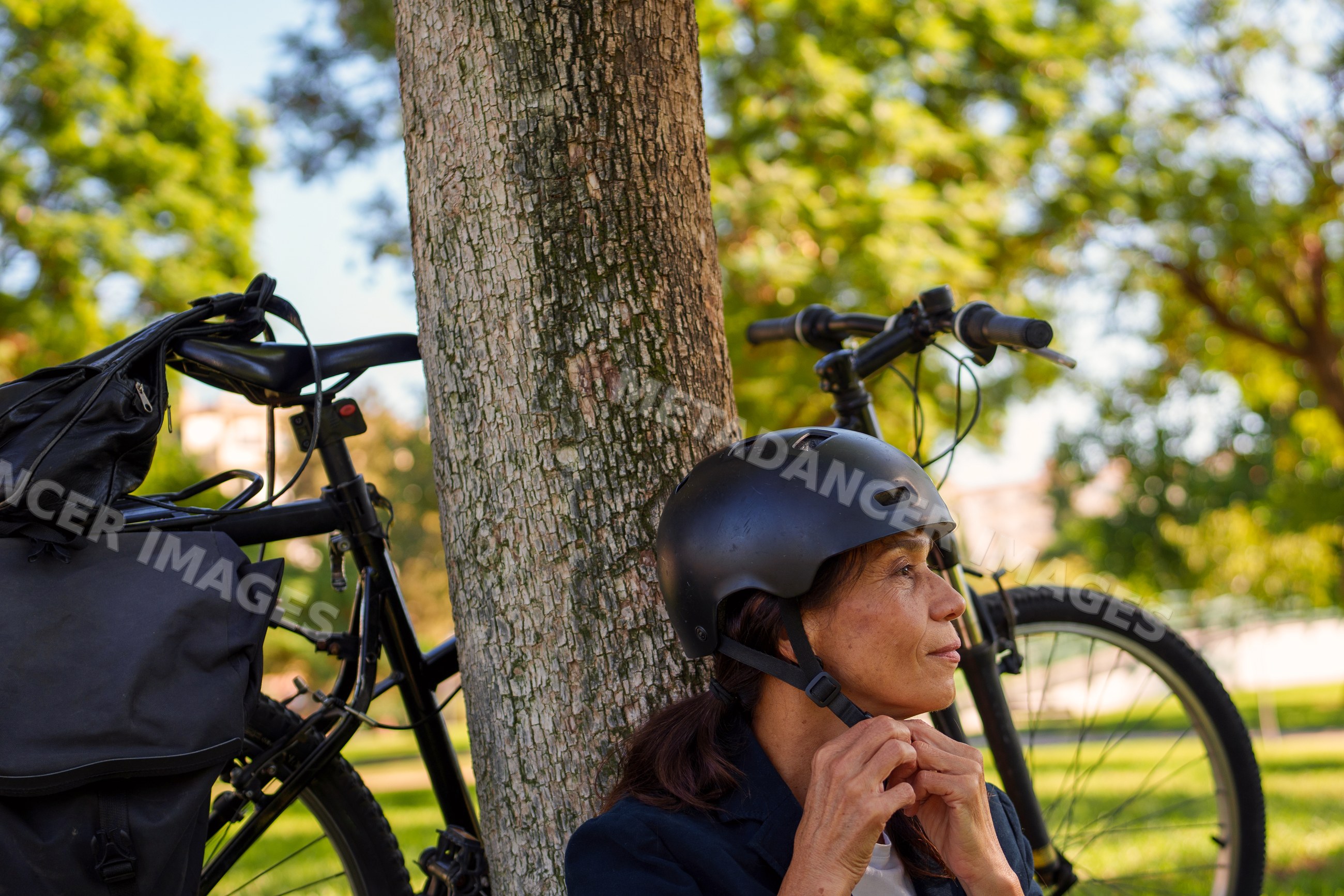 Hispanic woman putting on helmet by tree with bicycle in Málaga park