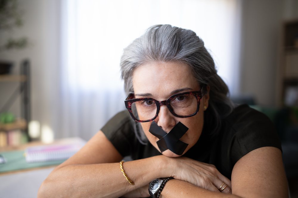 Older white woman with black tape on mouth resting arms indoors