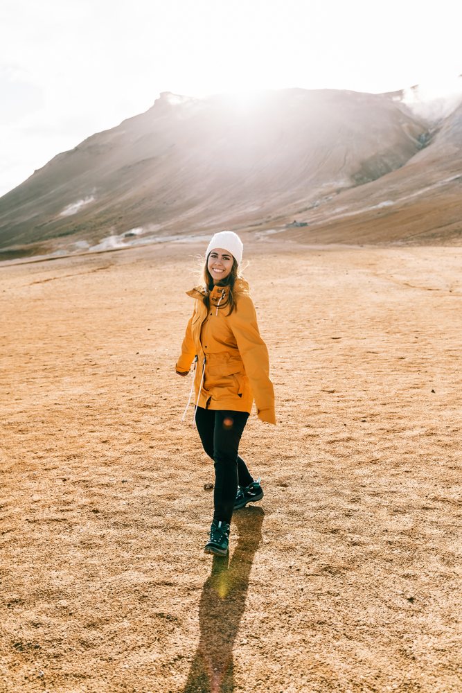 A white woman hiking in a yellow jacket enjoys a sunny mountain