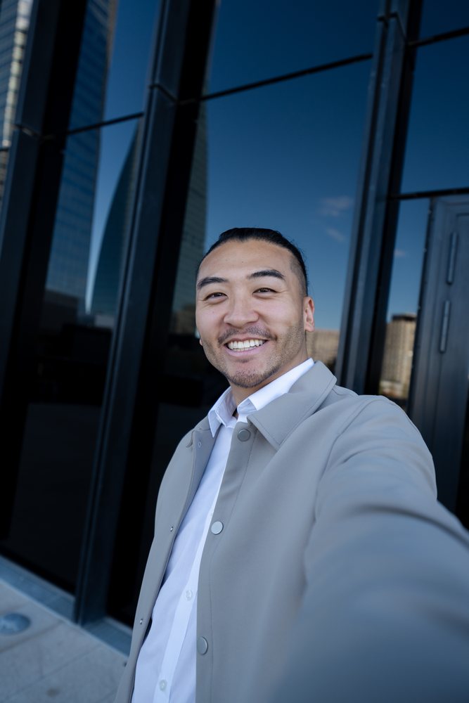 Man taking a smiling selfie outdoors by urban glass building