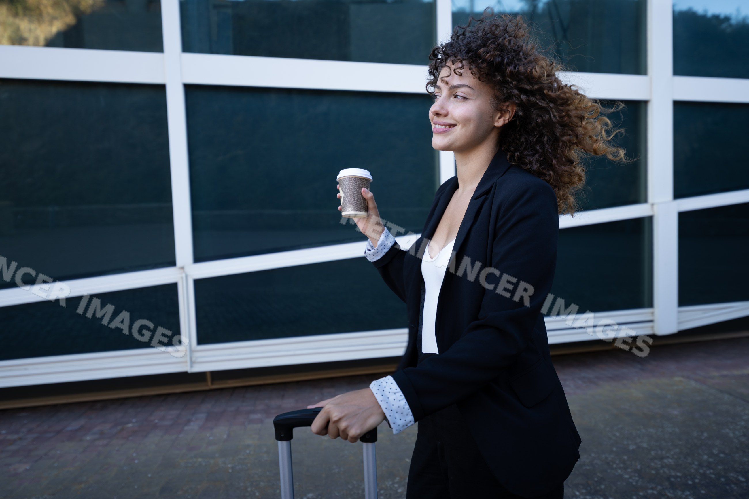 Businesswoman walking with coffee and suitcase by modern building