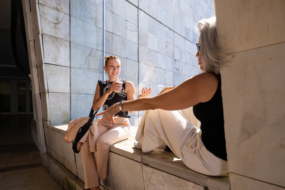 Two white women sitting and talking with coffee by window ledge