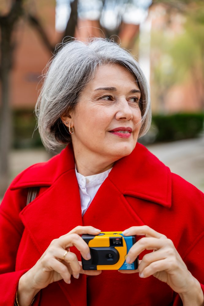 White woman holding disposable camera outdoors in red coat