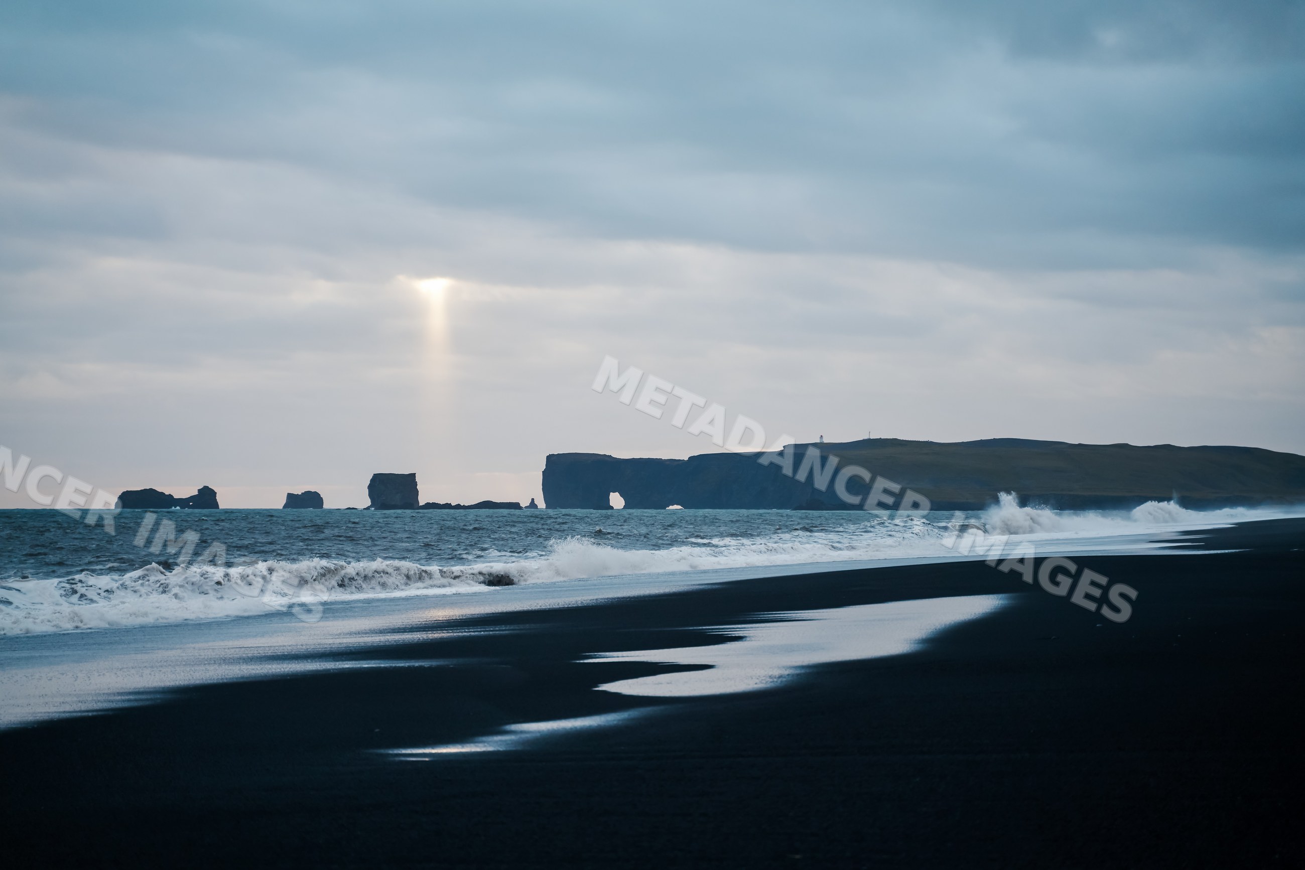 Waves crashing on a black sand beach with sea stacks under cloudy sky