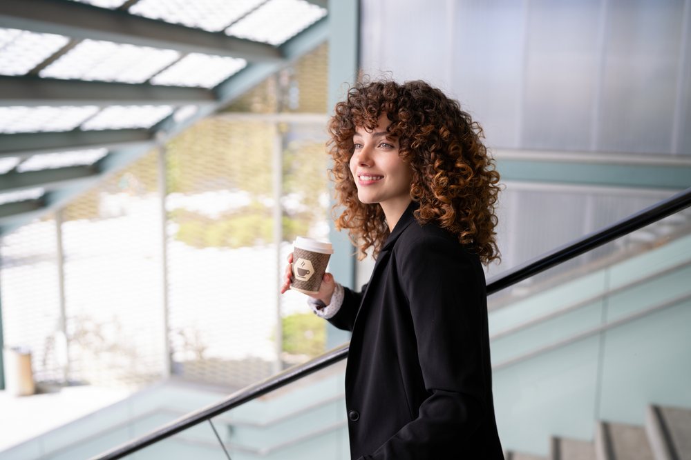 White woman with curly hair holding coffee on modern staircase