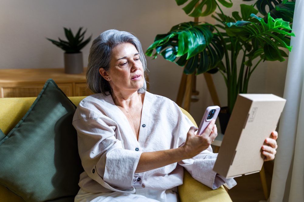 Middle-aged white woman unpacking box while using mobile phone indoors