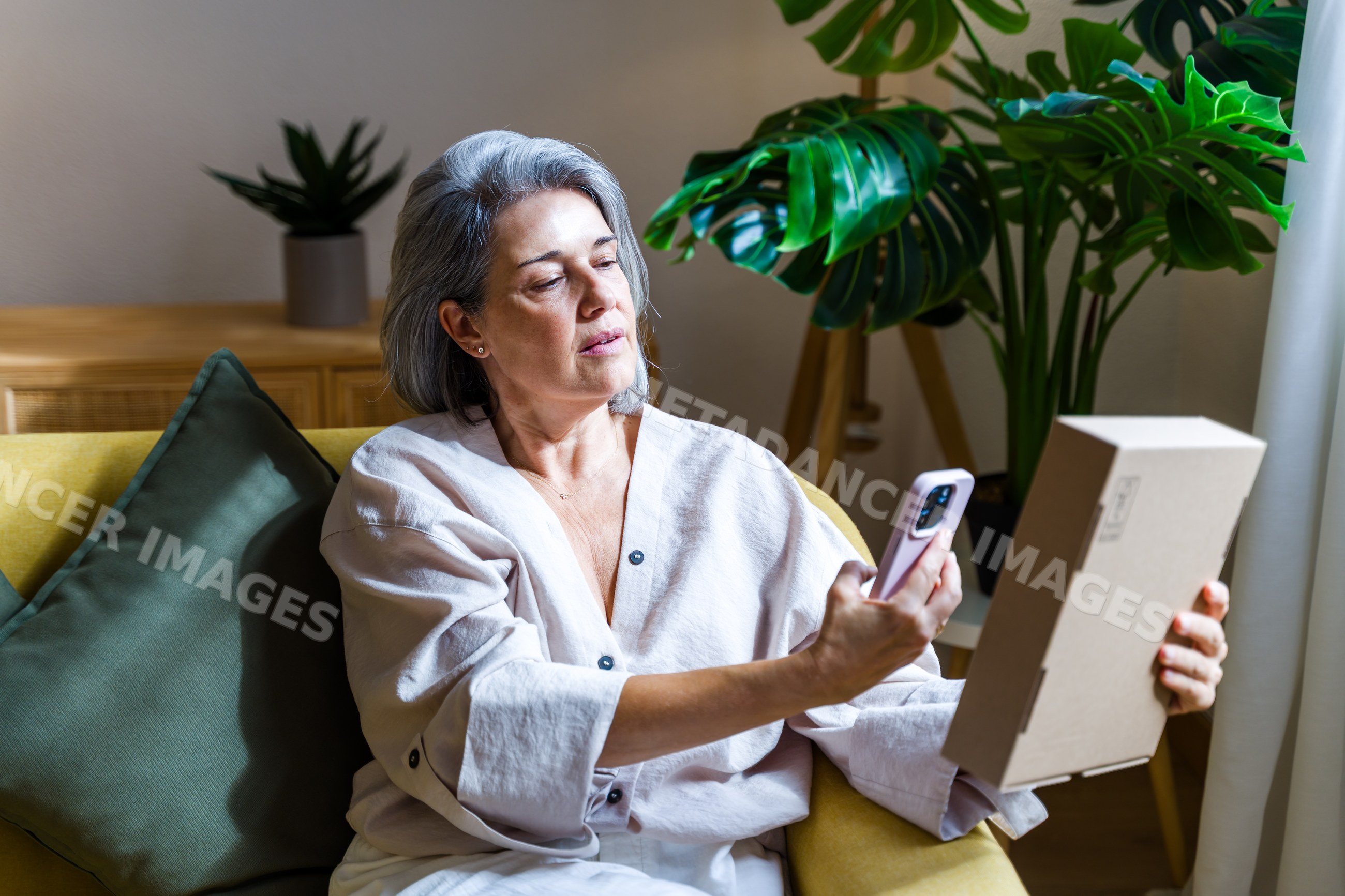 Middle-aged white woman unpacking box while using mobile phone indoors