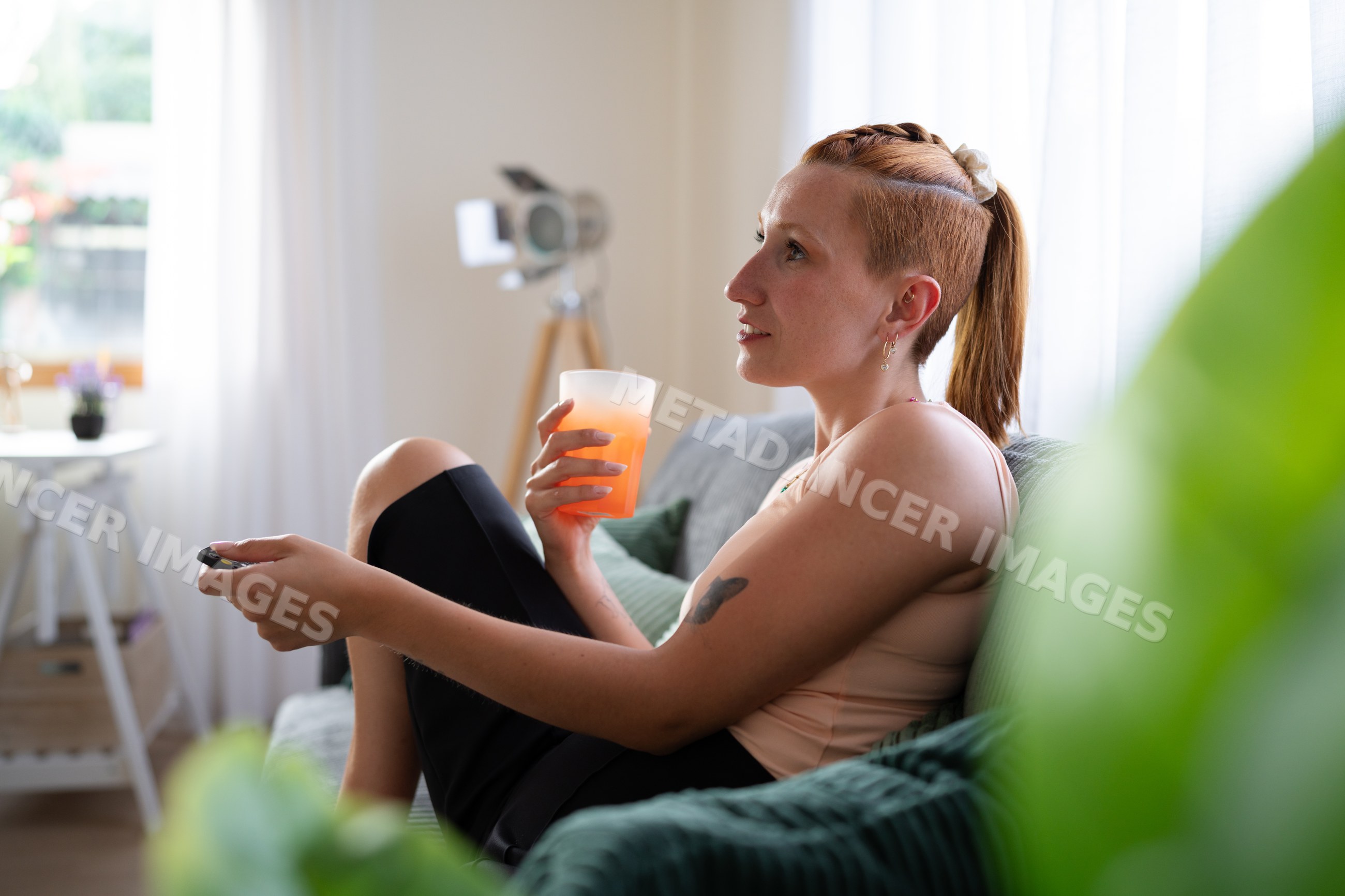 A white woman relaxing on couch with juice and remote control at home