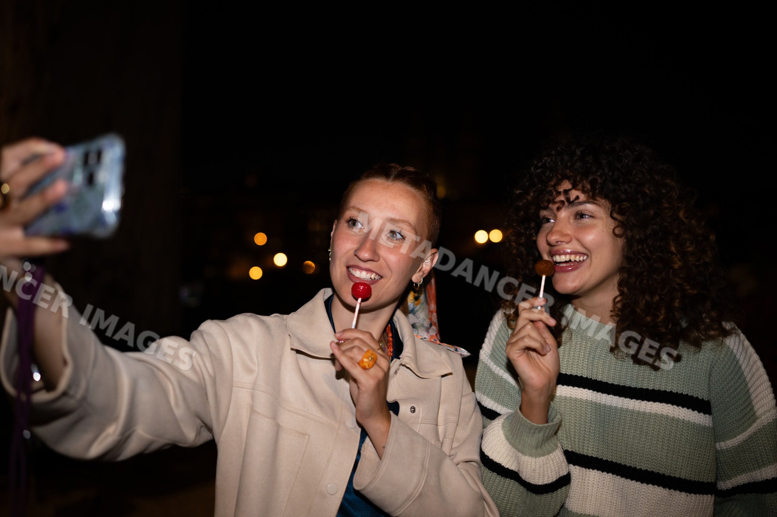 Two white women taking a selfie and holding lollipops at night