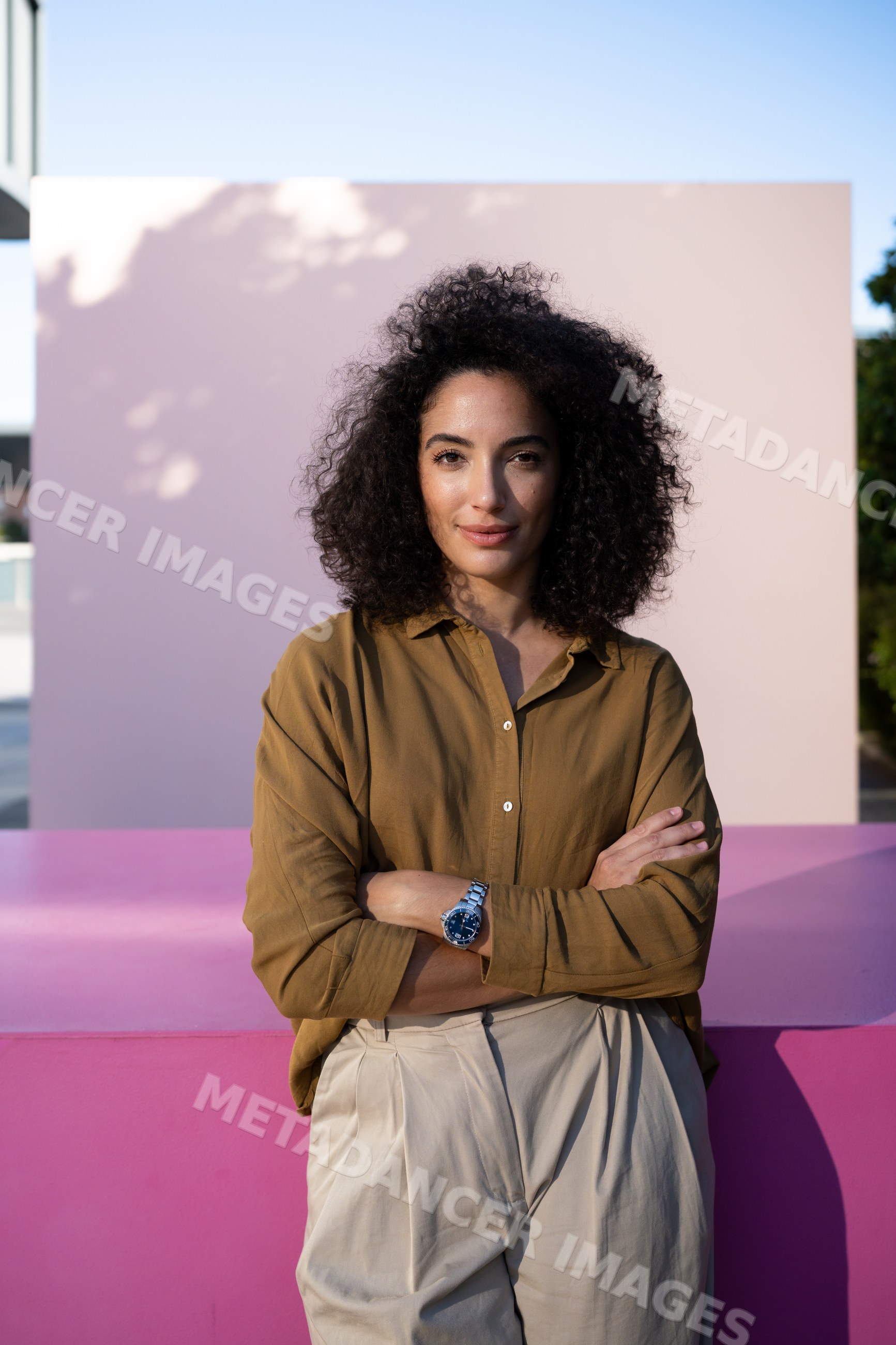 Woman standing with crossed arms against a pink wall outdoors