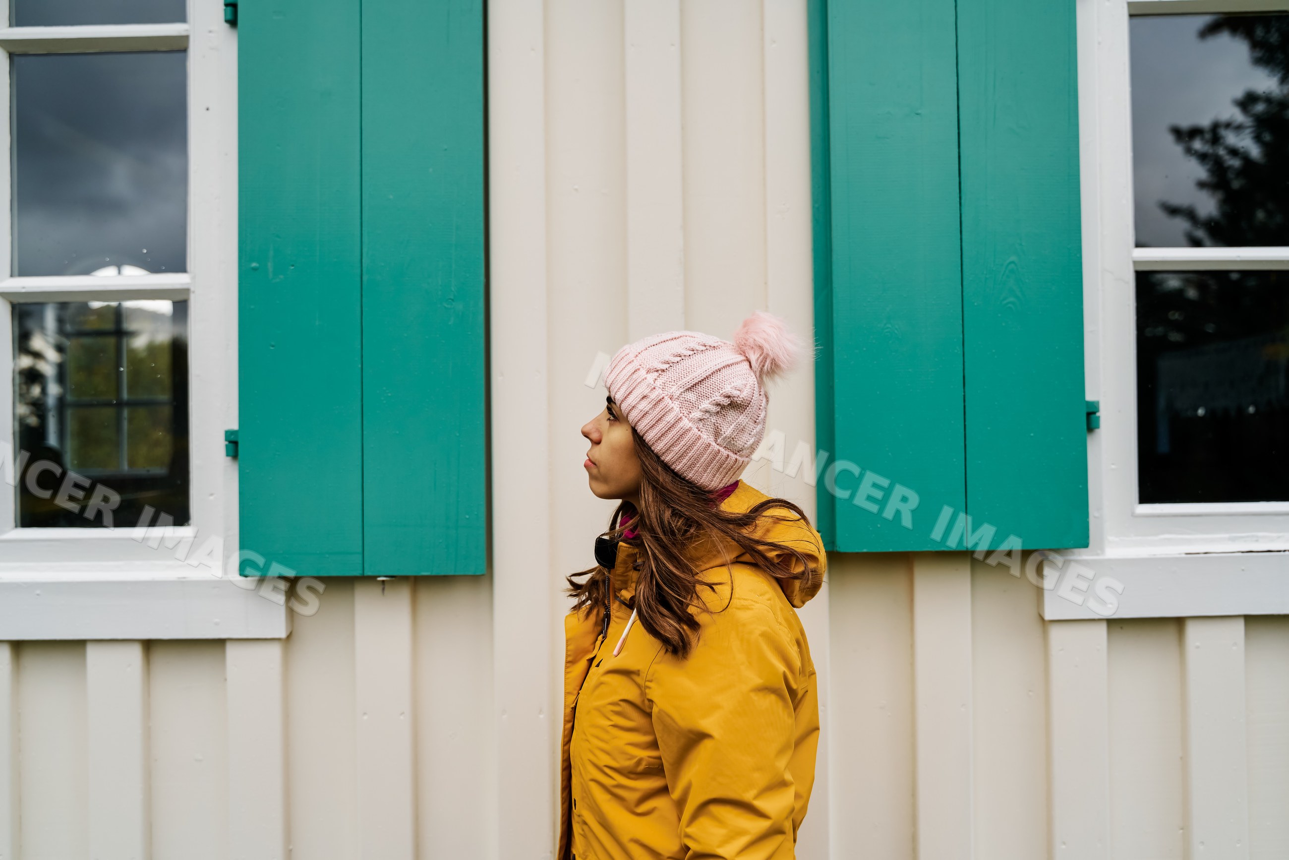 White Woman in yellow jacket and pink hat standing by a building