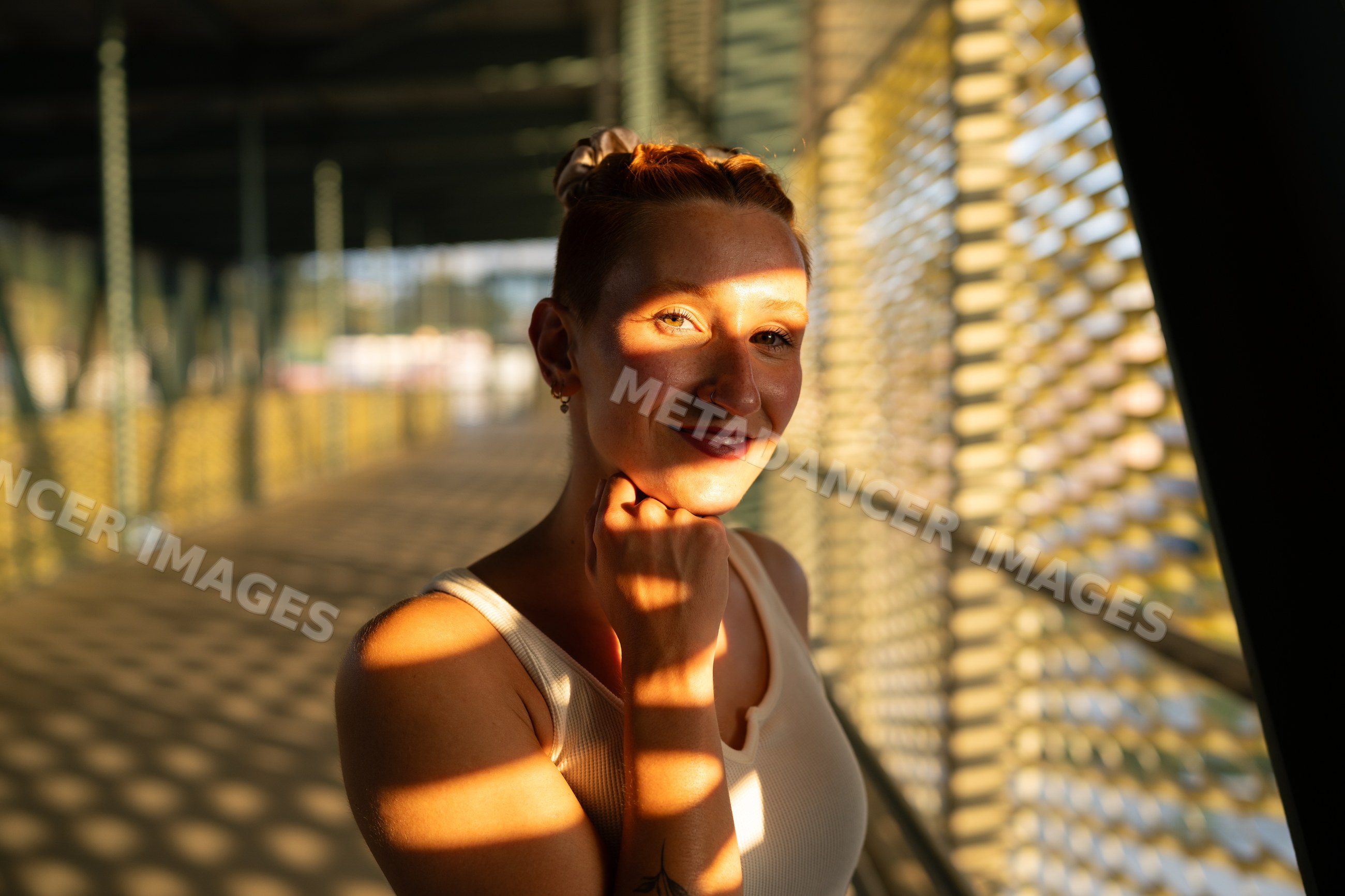 A white woman poses under patterned sunlight on a metal bridge walkway
