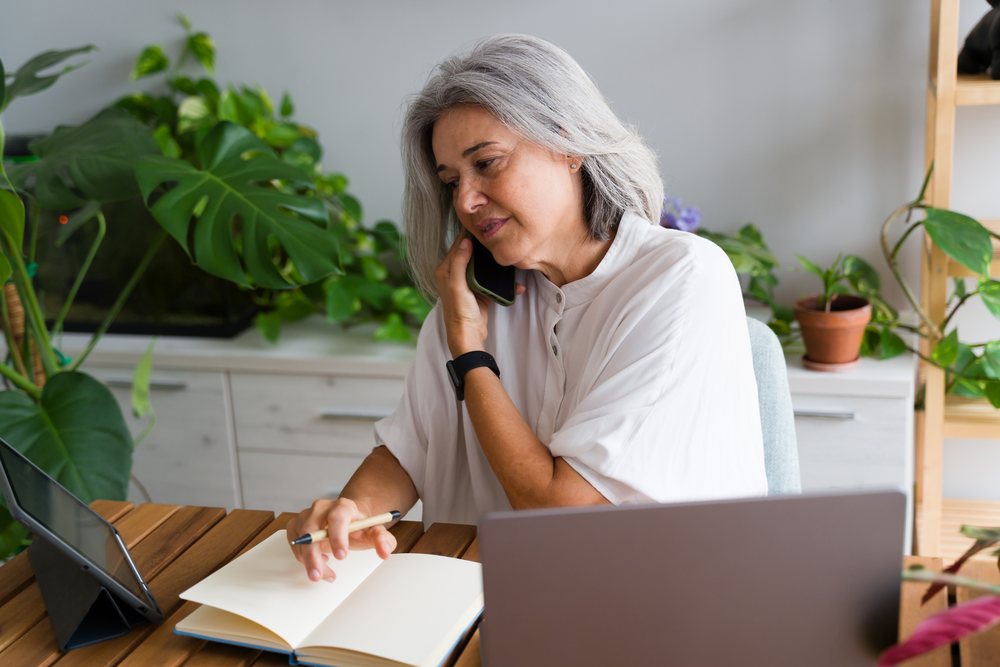 Mature white woman on phone working with notebook and laptop