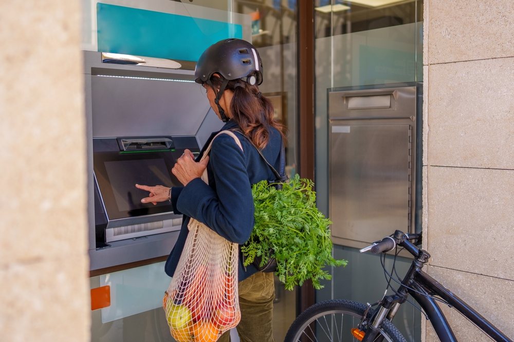 Hispanic woman withdrawing cash at outdoor atm carrying grocery bag
