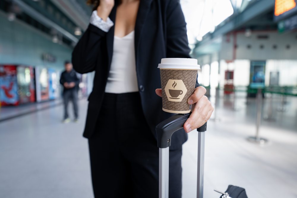 White woman holding coffee cup and luggage handle in airport terminal