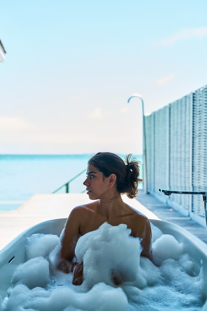 A white woman relaxes in a bubble bath with an ocean view