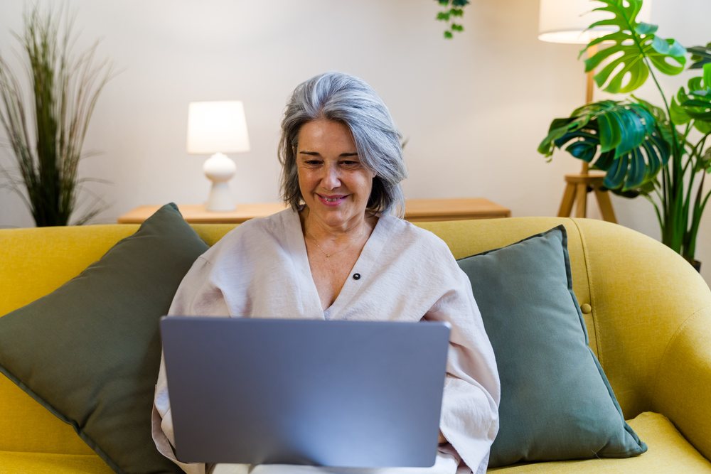 Senior white woman using laptop on yellow sofa in modern living room