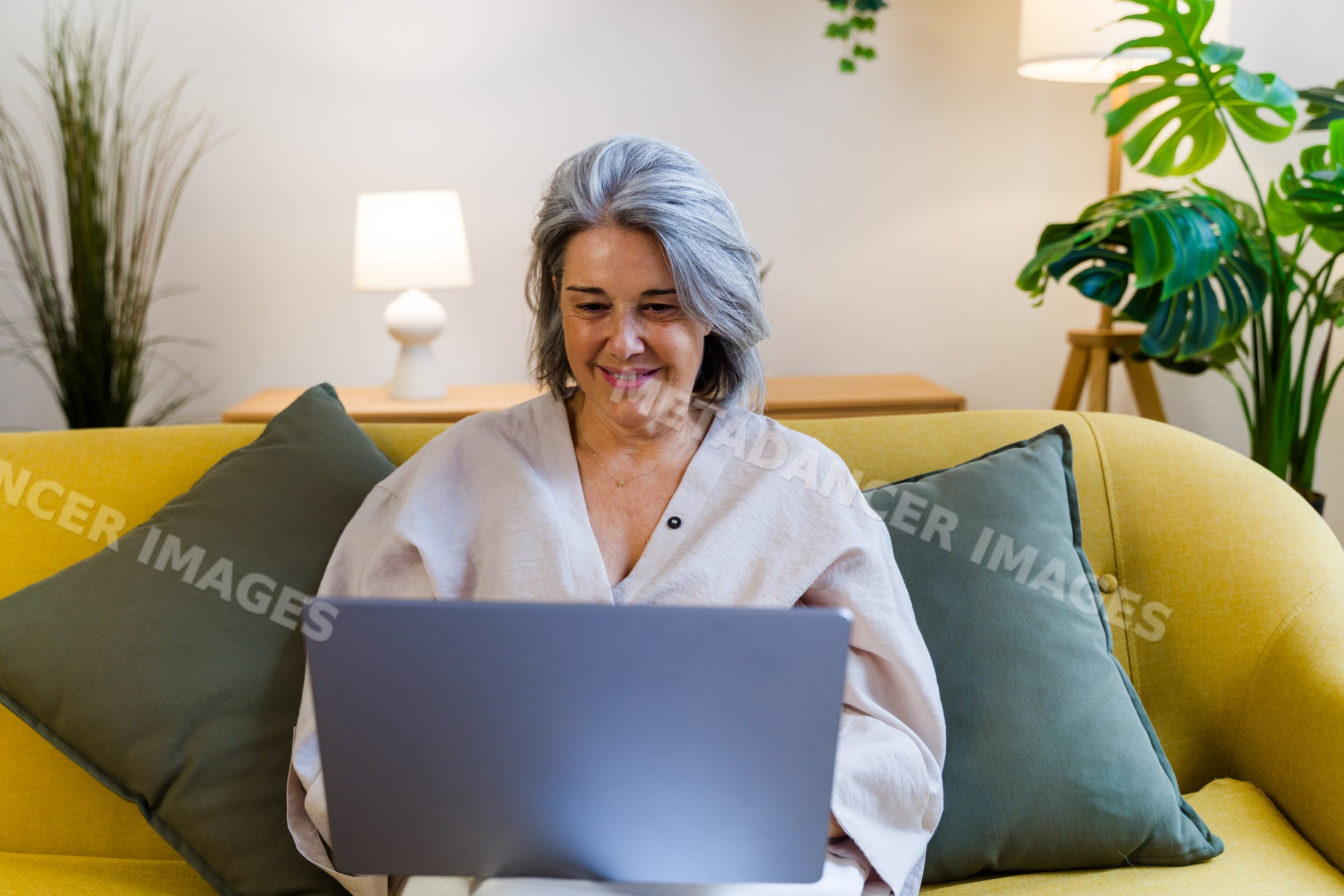 Senior white woman using laptop on yellow sofa in modern living room