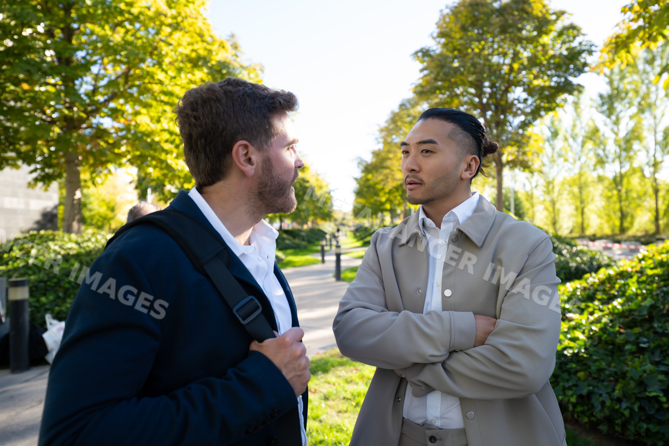 Two white men conversing on a sunny park pathway amid greenery