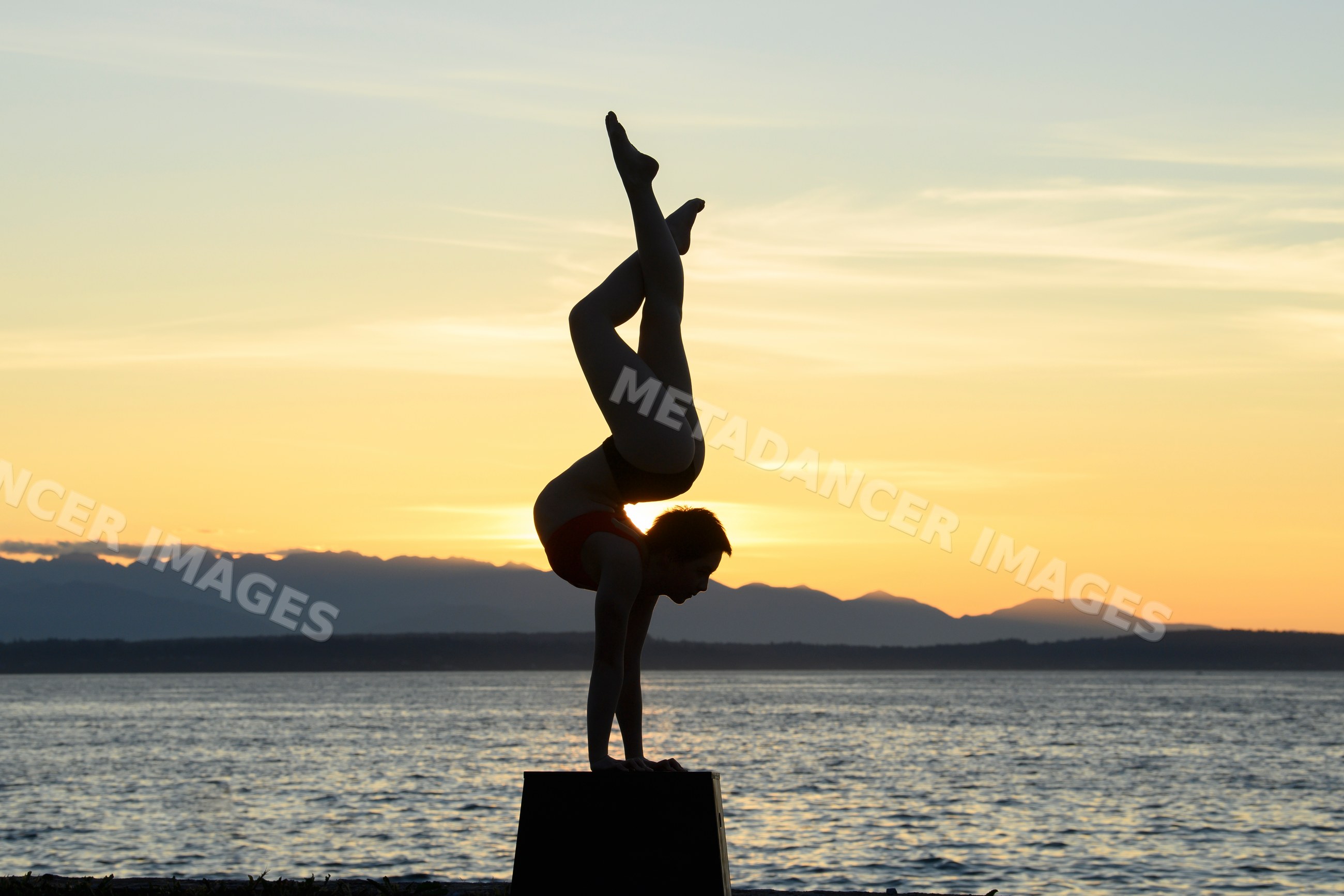 White woman doing handstand on box at ocean sunset with mountains