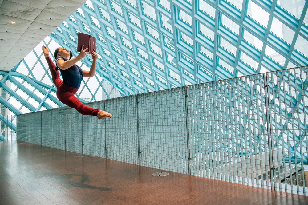 A woman jumps in a split leap holding a book in a library