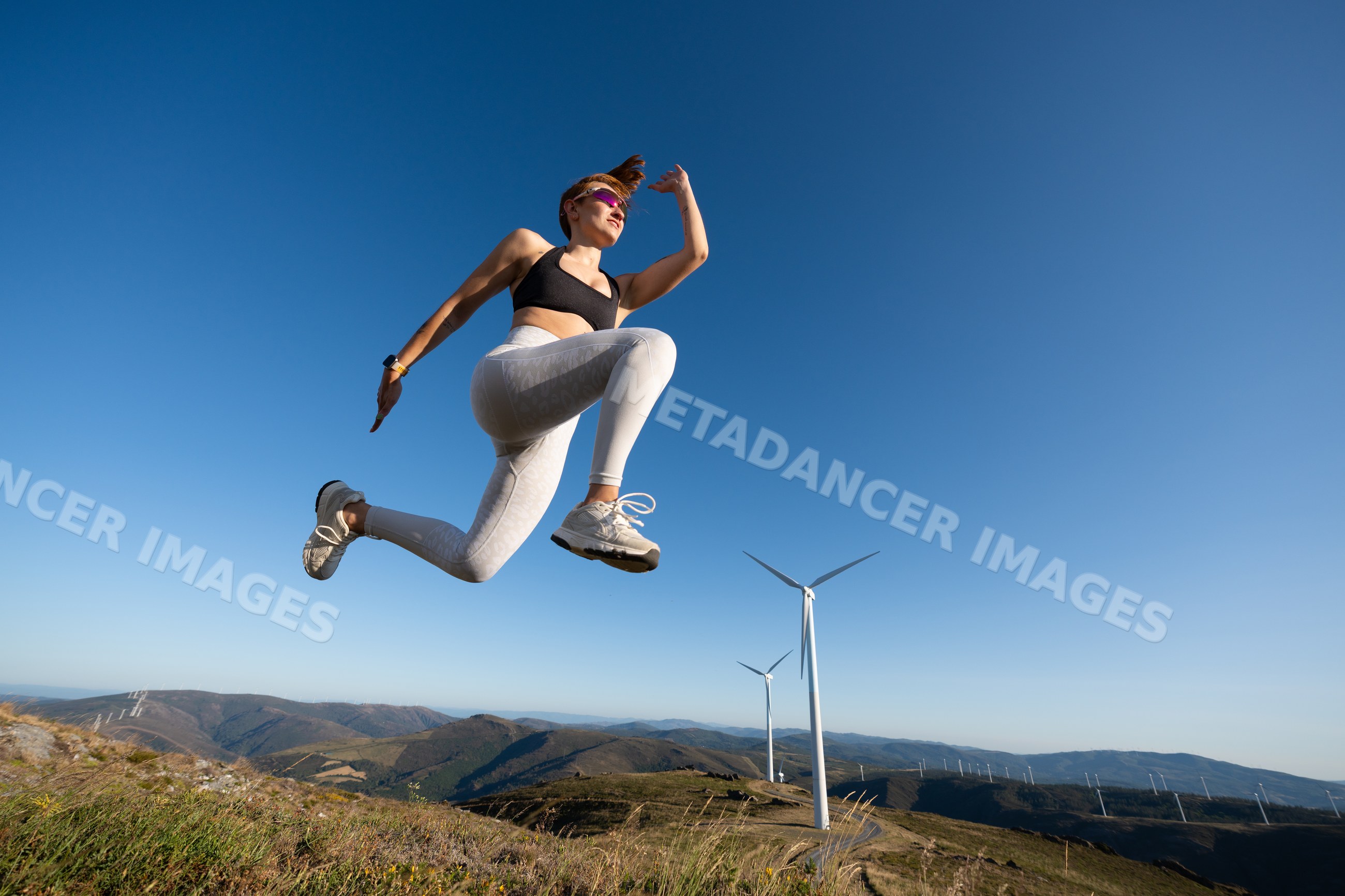 A white woman jumps on a hilltop near wind turbines in Galicia, Spain