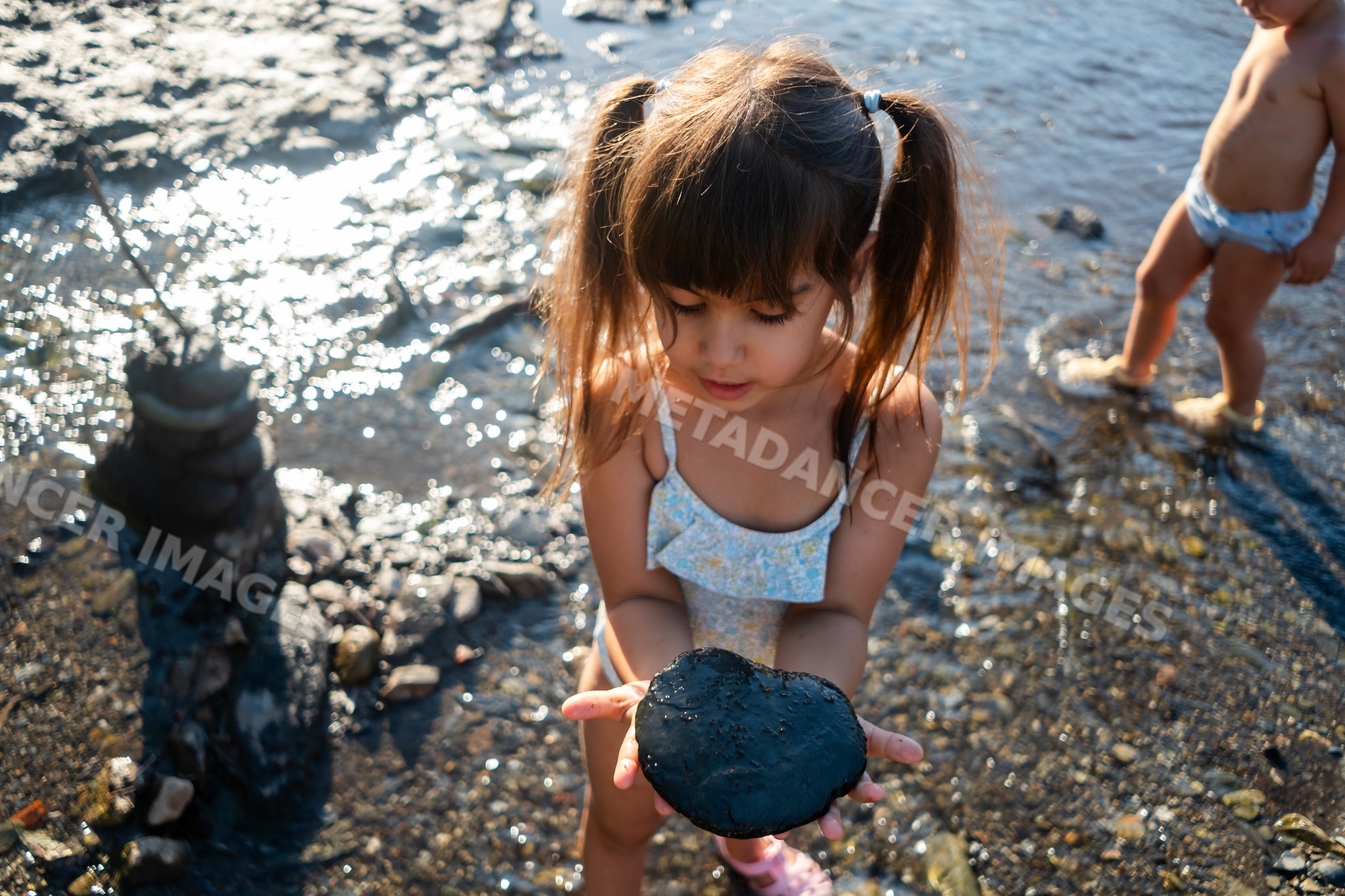 A young white girl holds a black rock by the water's edge